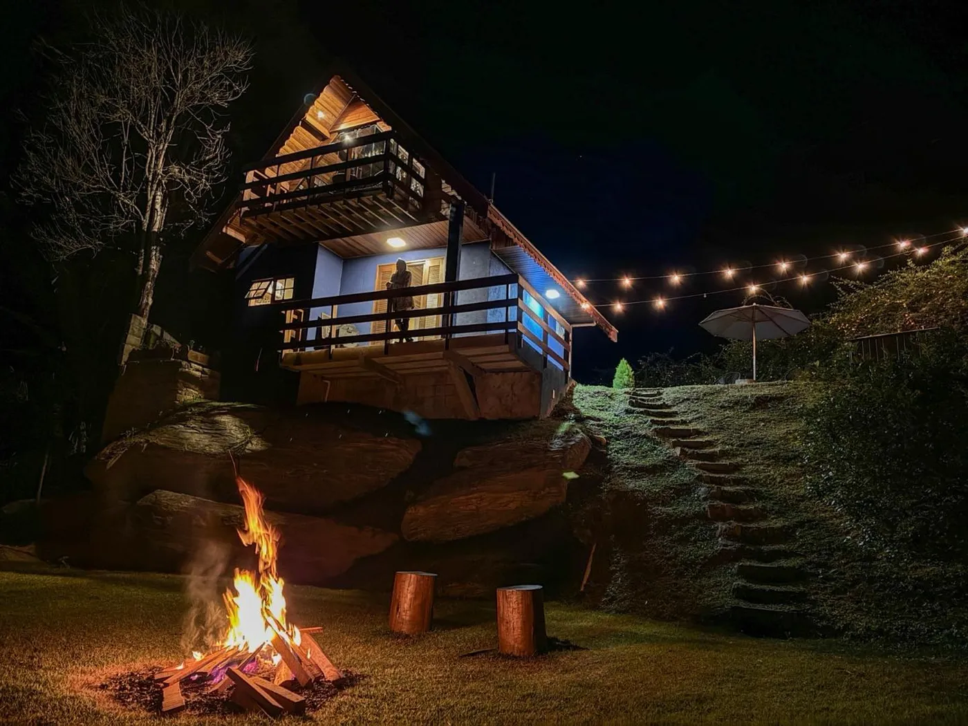 Fogueira acesa no jardim com o chalé iluminado ao fundo, à noite